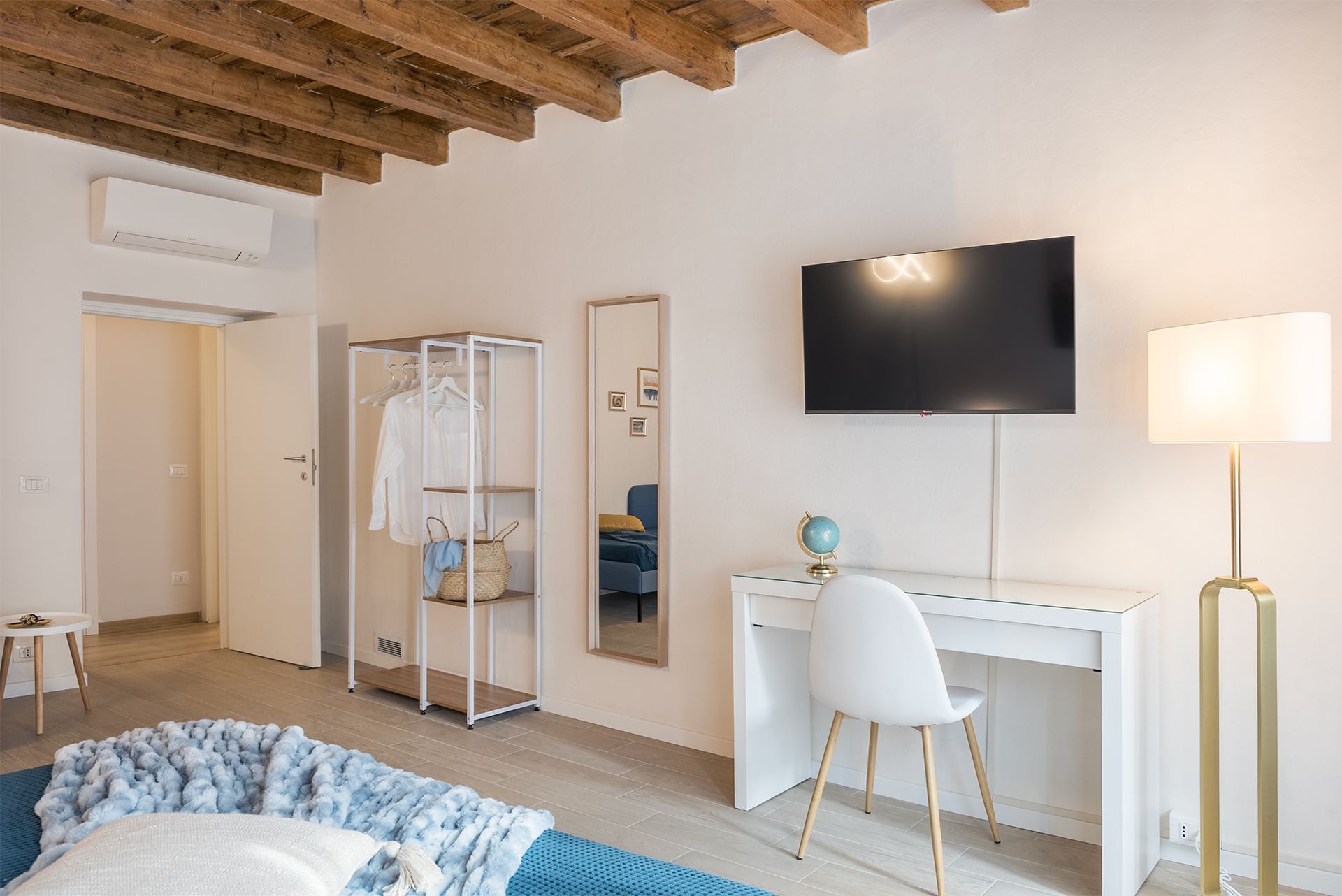 Another perspective of the bedroom with exposed beams, showing a desk with a white chair, floor lamp, and an open wardrobe.