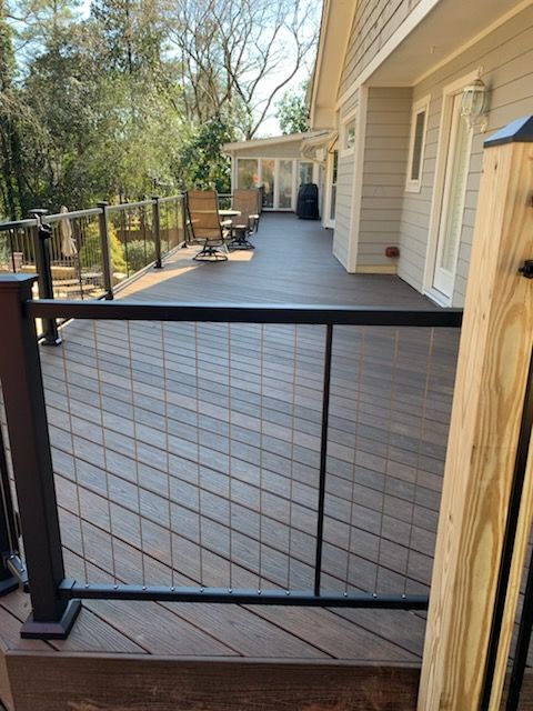 Black metal gate opening onto a gray wooden deck beside a beige house with a wooded yard beyond