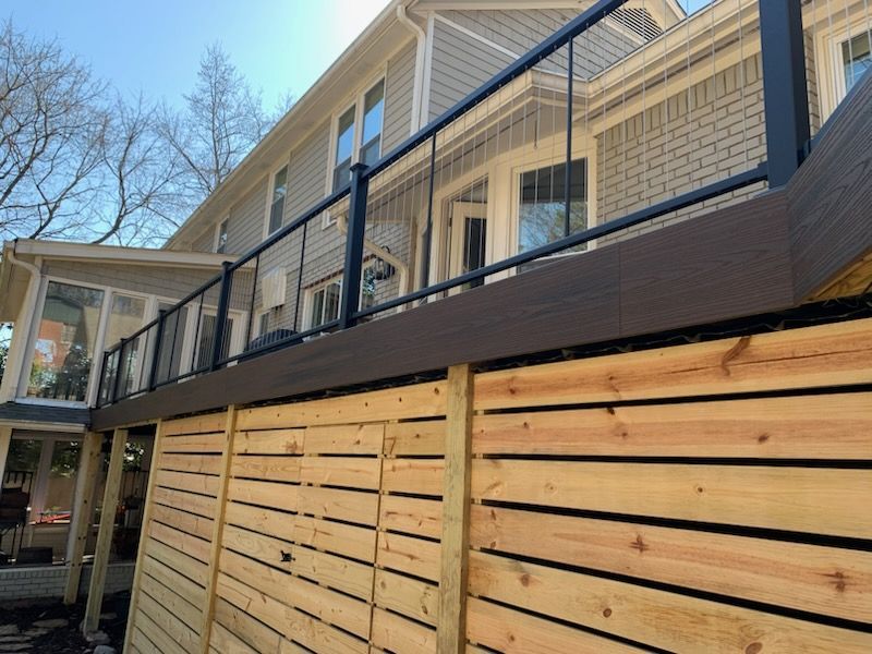 Wooden fence beneath a brick house deck with black railings on a sunny day