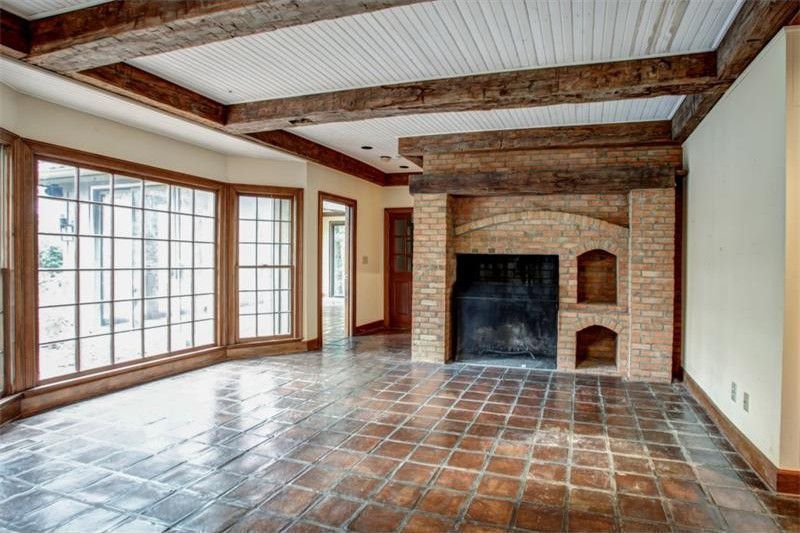 Empty living room with terracotta tile floor, brick fireplace, and large bay windows with wood beams