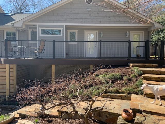 House with covered deck and stone steps in a sunny backyard, with a dog walking at right.