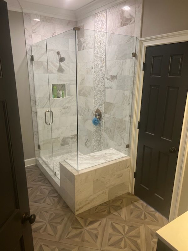 Glass walk-in shower with marble tile walls, stone bench, and dark door in a small bathroom