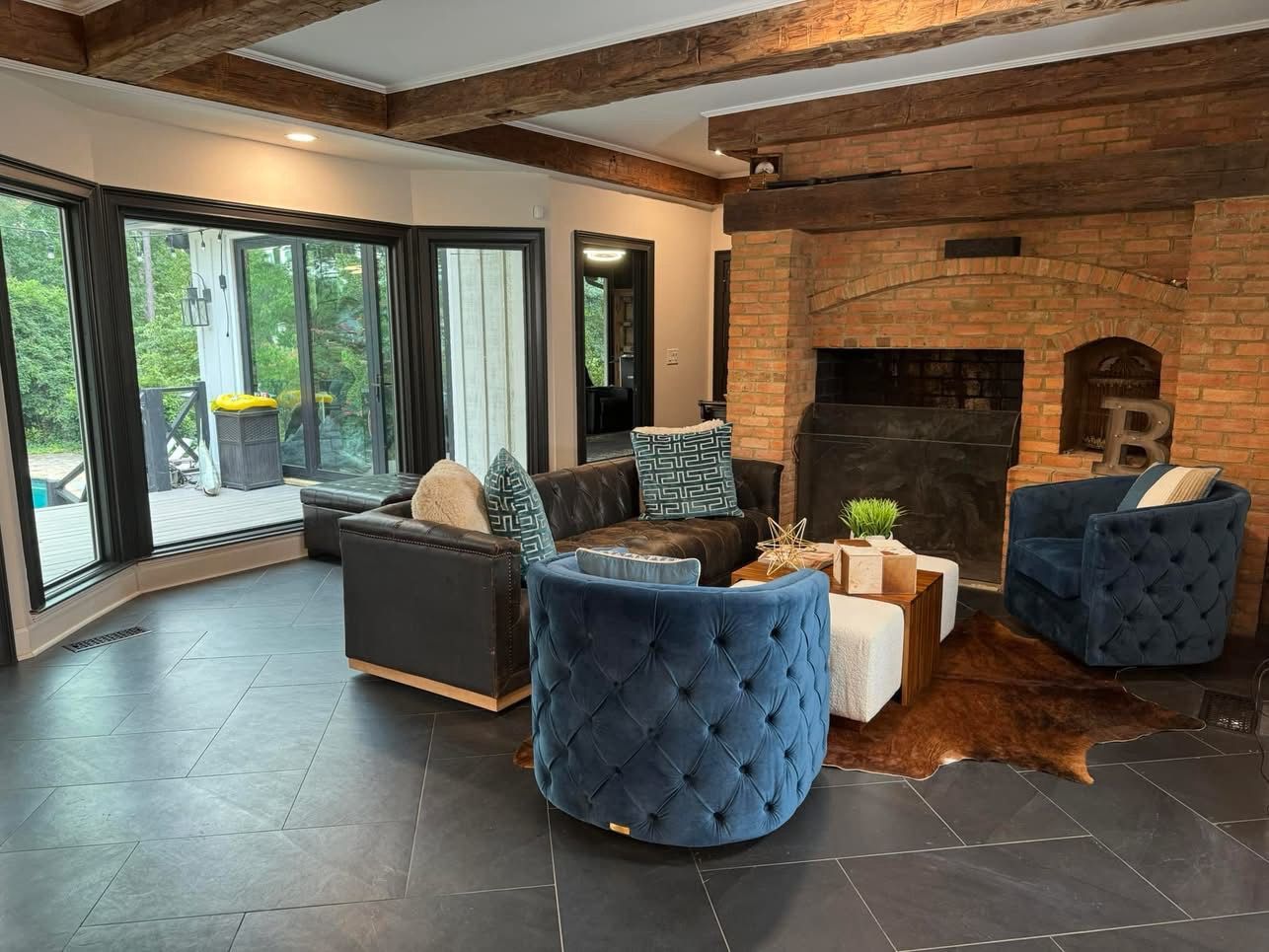 Modern living room with blue chairs, dark sofa, large windows, and a brick fireplace