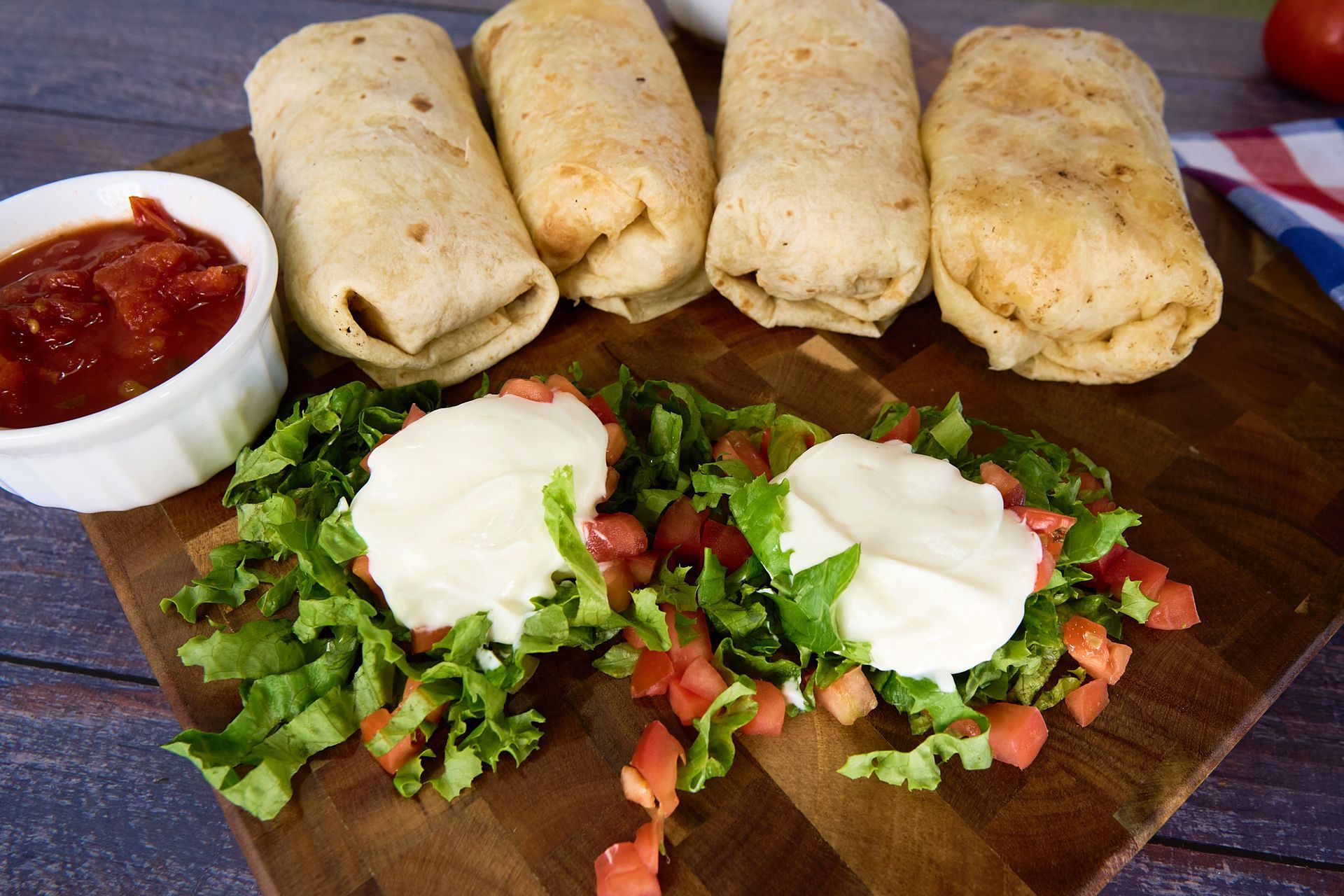 A wooden cutting board topped with burritos , lettuce , tomatoes and sour cream.