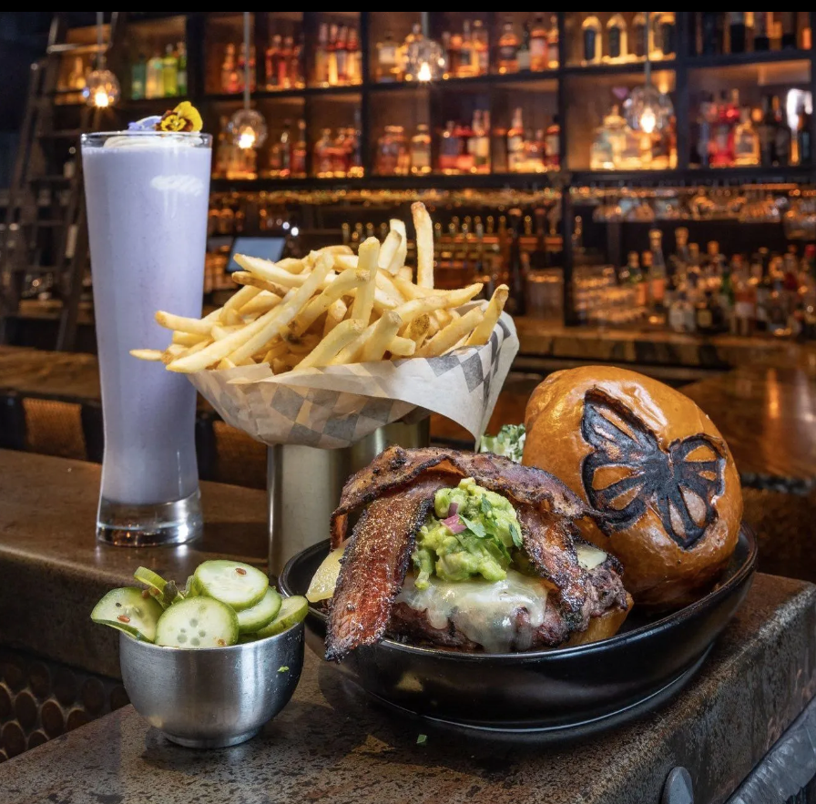A hamburger , french fries , bacon and a milkshake are on a table in a restaurant.