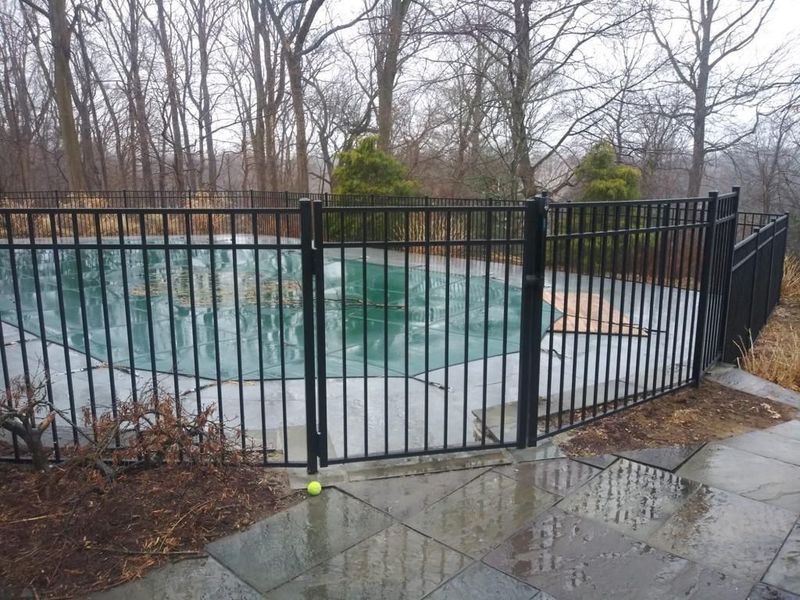A black fence surrounds a swimming pool with trees in the background