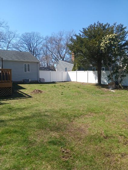 A backyard with a white fence, grass, and a tree. A house is on the left. Sunny day.