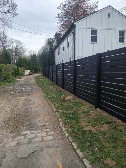 Black horizontal slat fence alongside a gravel driveway, next to a white house. Cloudy sky.