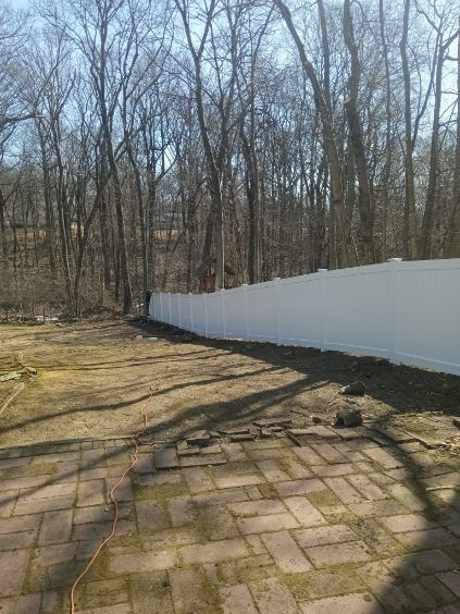 A long, white vinyl fence borders a backyard with a brick patio in the foreground and a wooded area beyond.