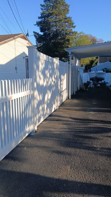 White fence lines a driveway, with a car port and house visible. Sun casts shadows.