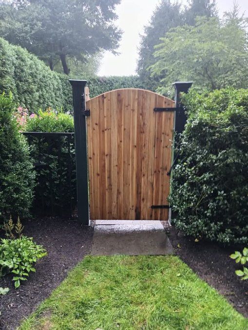 Wooden gate with arched top, flanked by green hedges, leading into a garden path.