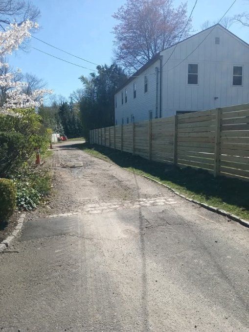Paved driveway next to a new wooden fence, leading towards a white two-story building on a sunny day.