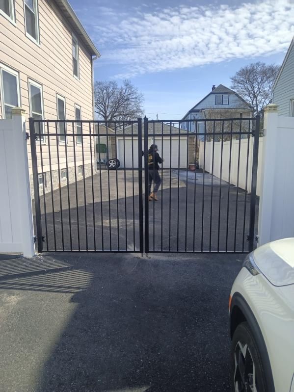 A black metal gate opens onto a paved driveway. A person stands near the gate. Buildings frame the scene under a partly cloudy sky.