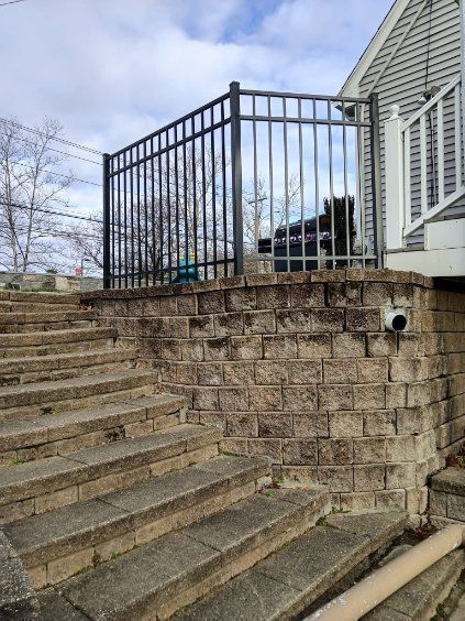 Outdoor stone stairs lead to a retaining wall with a black metal fence and house. Cloudy sky.