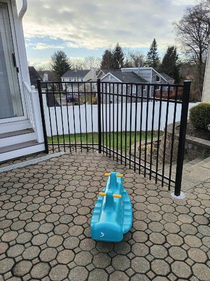 A blue rocking horse on a brick patio, fenced with black metal, houses visible in the background.