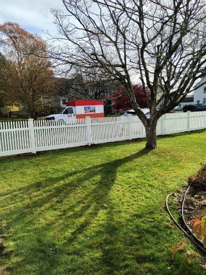 White picket fence with a tree casting a shadow on green grass, white moving truck in the background.