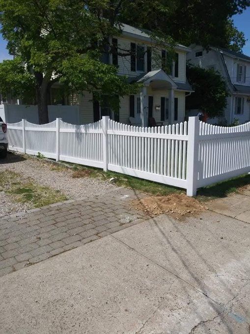 White picket fence in front of a two-story house, gravel driveway, and brick sidewalk.