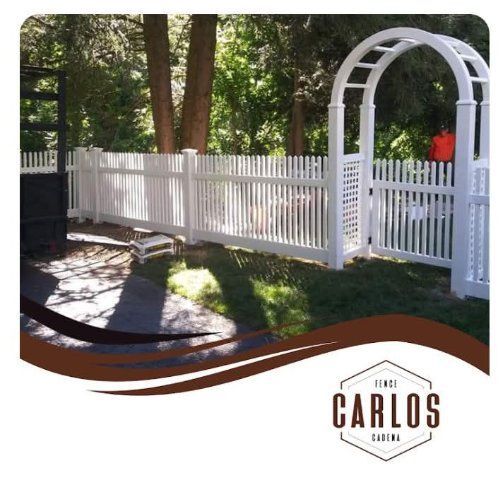 White picket fence with archway in a grassy yard, trees in the background, company logo in bottom corner.