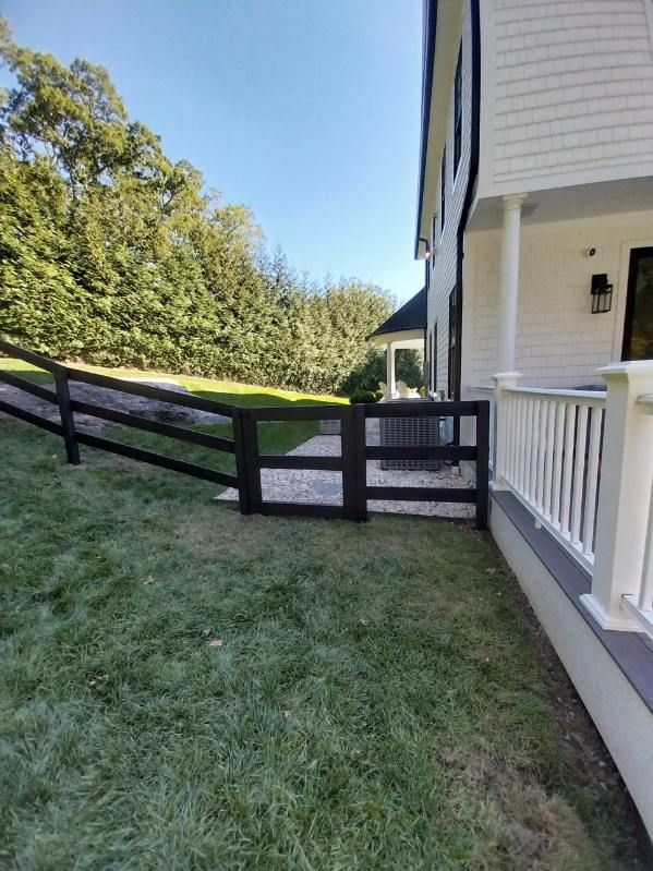 Black wooden fence near a white house and porch, with green grass and a gravel area.