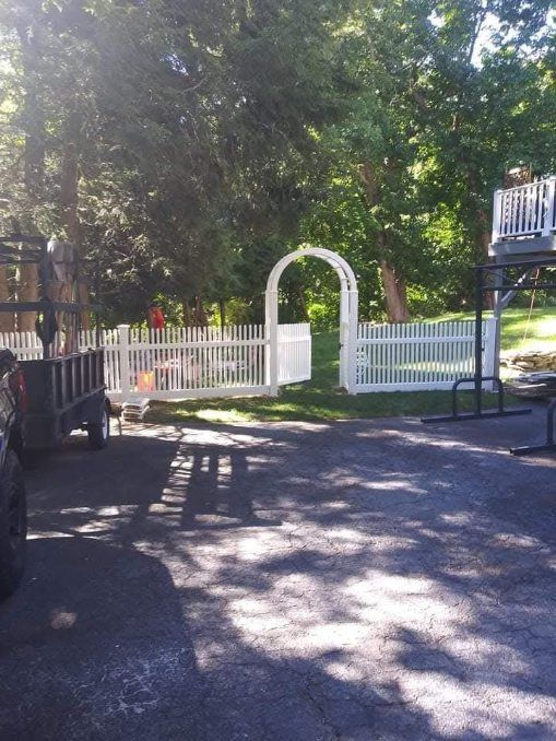 White picket fence with an archway, trees in the background, set on a driveway.