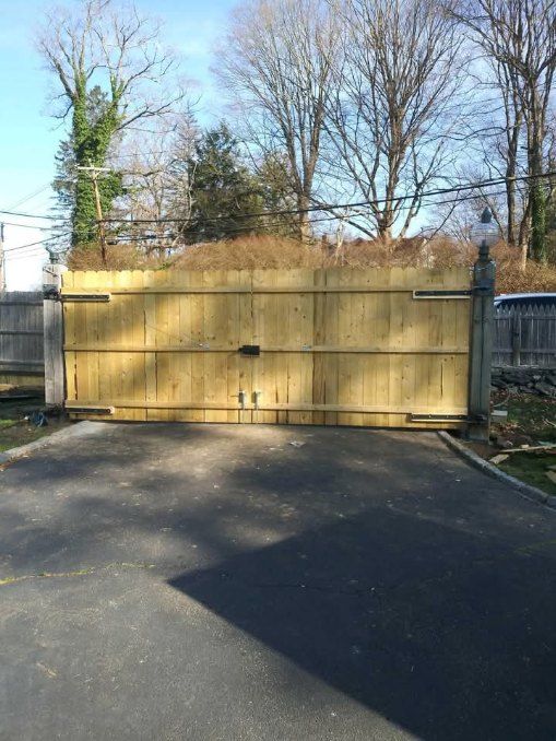 Wooden driveway gate, closed, on asphalt driveway. Fence and trees in background.