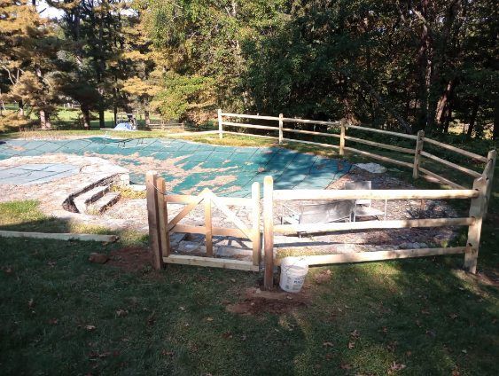 Wooden fence surrounds a pool covered with a green tarp, with a gate, in a grassy yard.