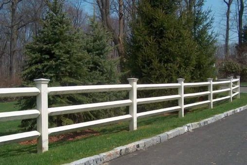 White three-rail fence alongside a paved driveway, with green grass and trees in the background.