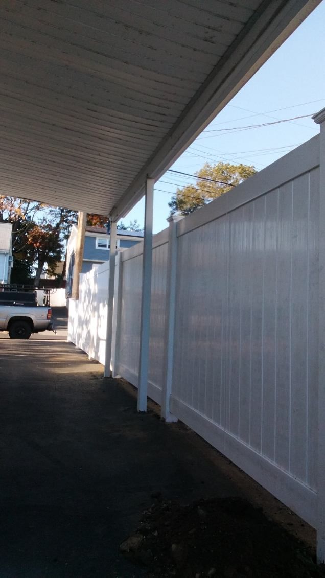 White vinyl fence alongside a covered driveway. Sunlight illuminates the scene, sky in the background.