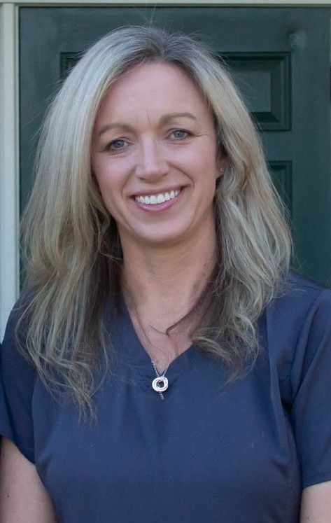 A woman in a blue scrub top is smiling in front of a door.