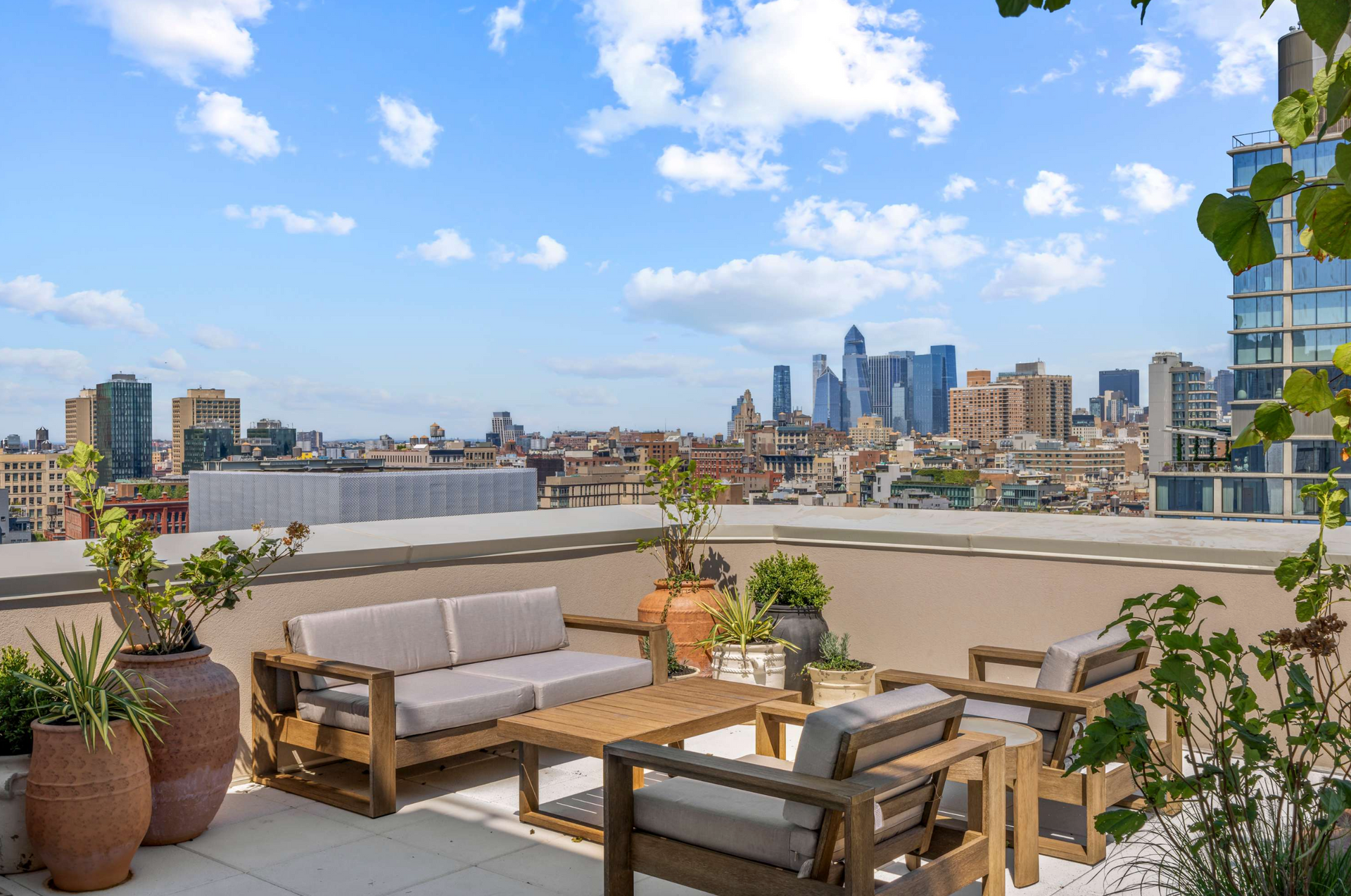 Rooftop patio with wooden furniture, overlooking a cityscape under a blue sky with clouds.