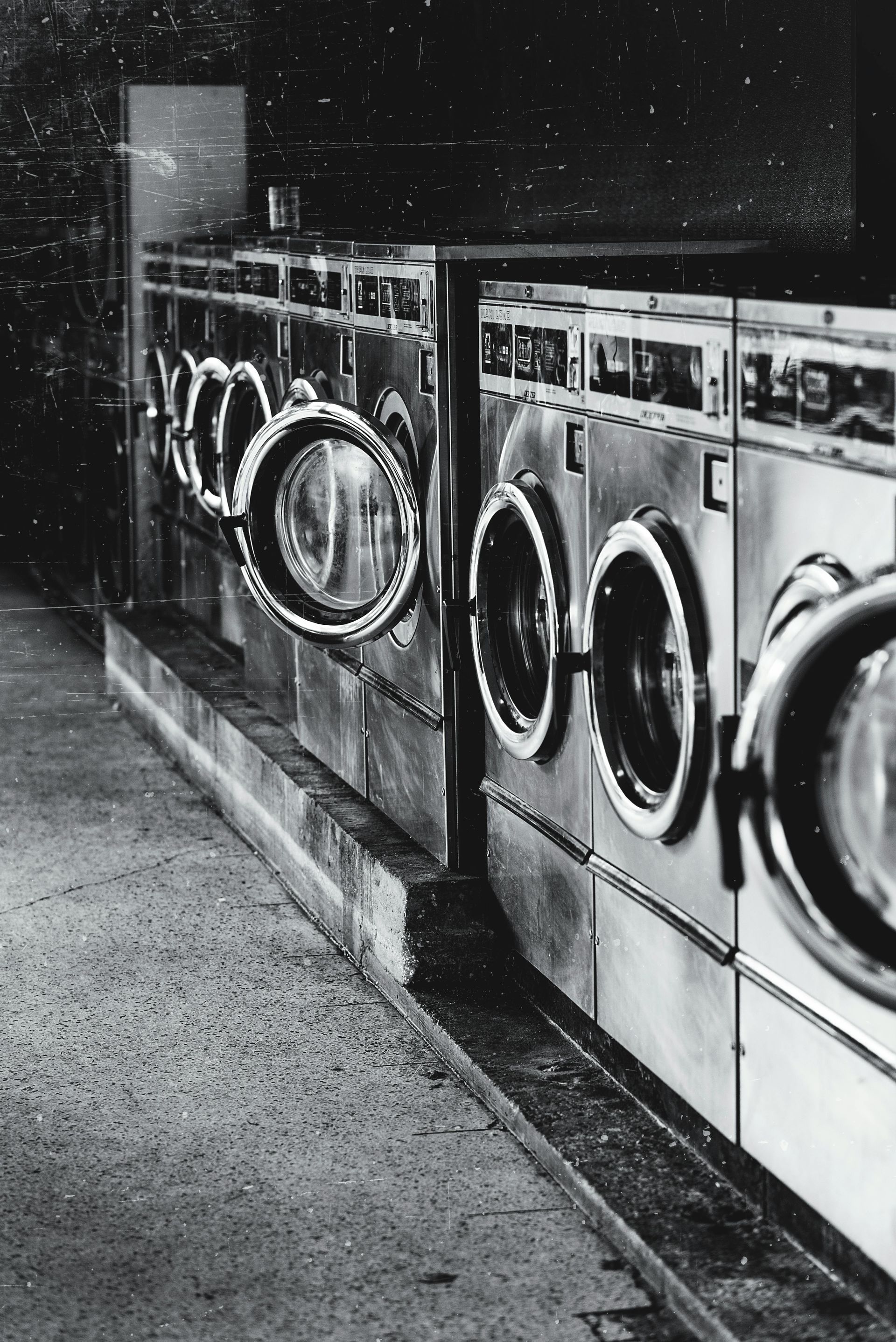 Row of washing machines in a laundromat, mostly silver with glass doors. Black and white.