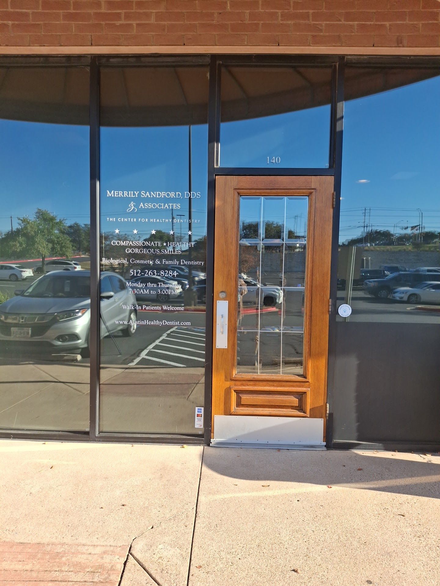 Storefront with glass windows and a wooden door, reflecting parked cars in a sunny shopping center.