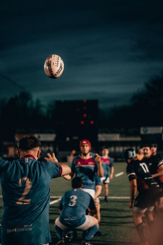 Rugby scrum under stadium lights, with the ball arcing overhead as players in dark and teal jerseys brace for play