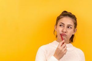 Person in a white sweater holding a red pencil and looking thoughtfully against a bright yellow background