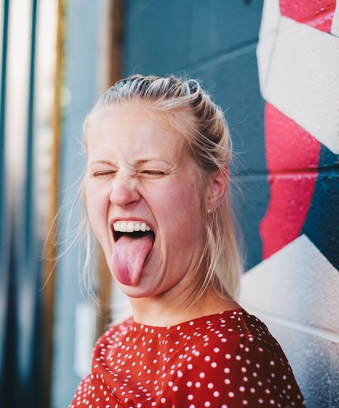 Smiling woman in a red polka-dot shirt sticking out her tongue beside a colorful wall