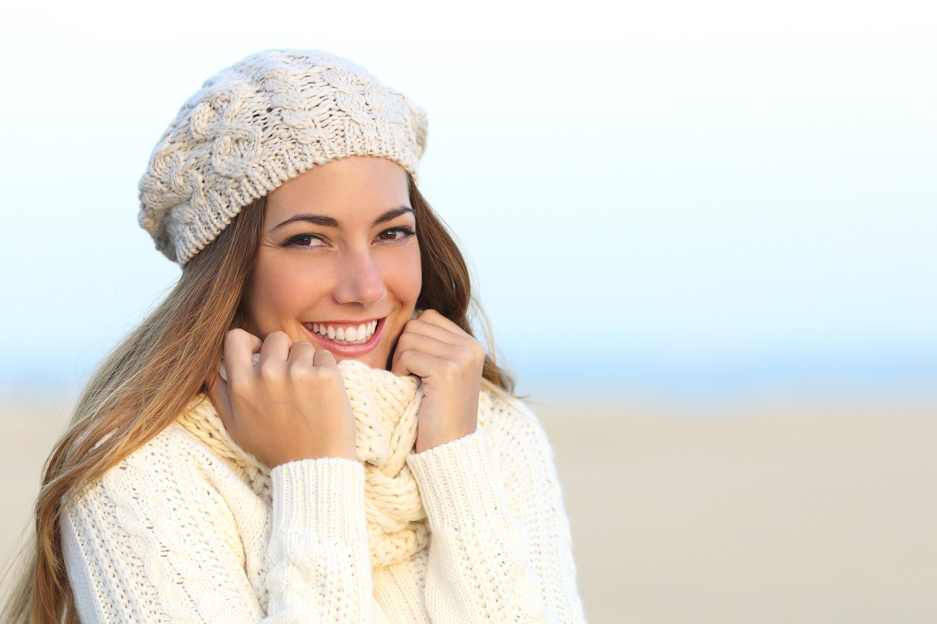 Smiling person in a cream sweater and knit hat outdoors on a beach, holding their collar in the wind
