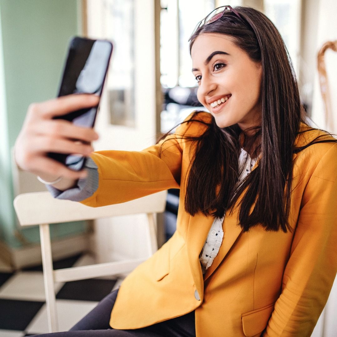 Woman in a yellow blazer taking a selfie indoors and smiling at her phone