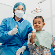 Dentist and young patient in blue scrubs and bibs giving thumbs up in a bright clinic