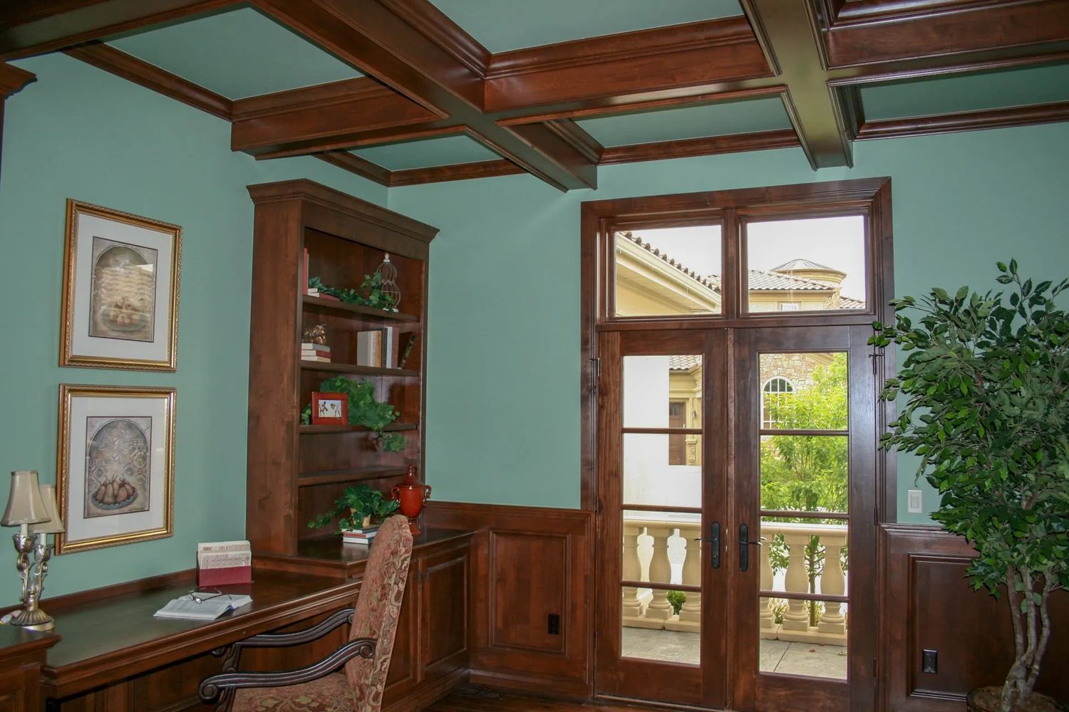 A wood-paneled office with a desk, bookshelf, and double doors leading to a balcony. Aqua walls and a plant.