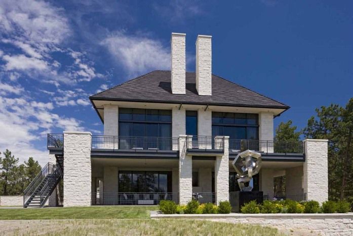 Modern two-story house with stone facade, large windows, and black metal railings. Clear blue sky.