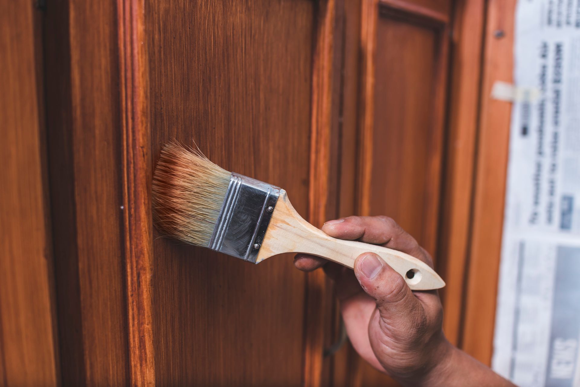 Hand holding a paintbrush applying brown stain to a wooden door panel