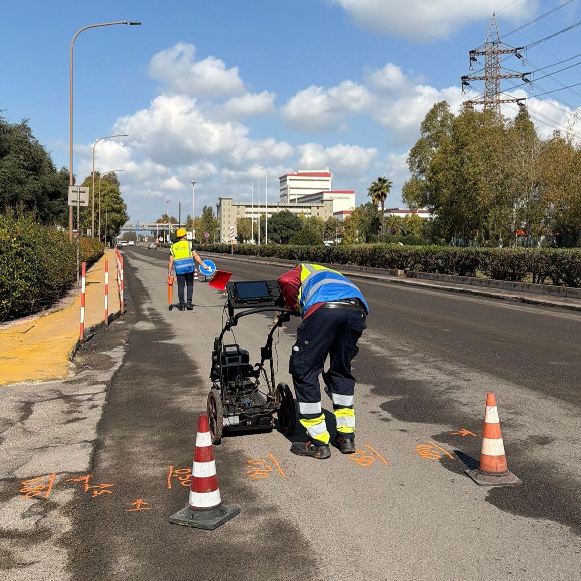 Operai stradali che segnalano la pavimentazione su una strada soleggiata, con coni, un carrello e una persona con un giubbotto di sicurezza blu.