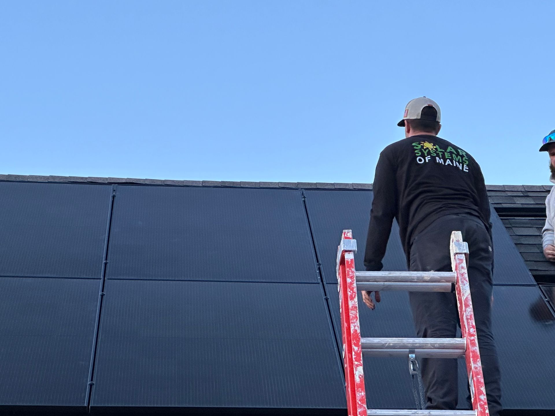 Person on a ladder installing solar panels on a rooftop against a clear blue sky.