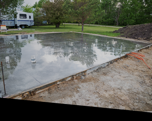 Wet concrete slab reflecting trees, with an RV in the background and an orange hose at the edge.
