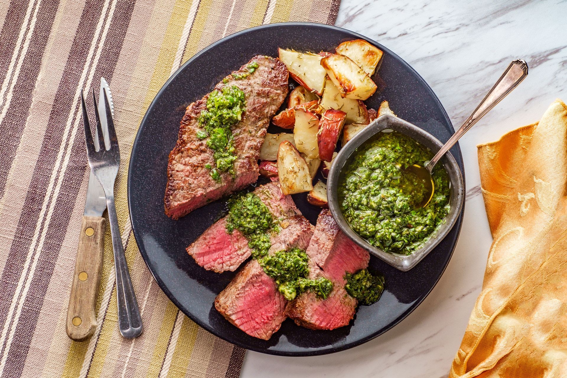 Medium-rare chimichurri steak with roasted potatoes and fresh herb sauce served on a dark plate.