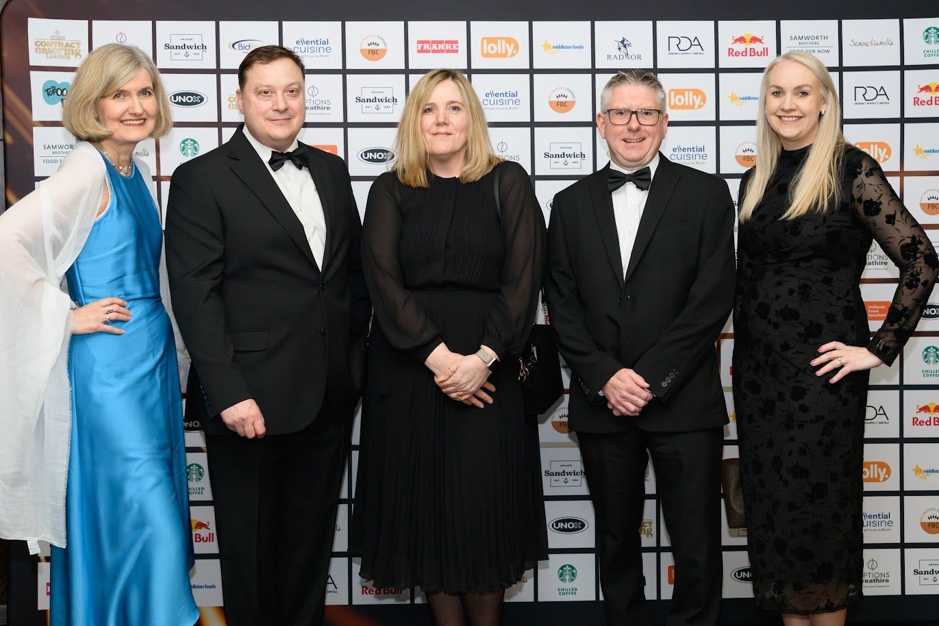 A group of five people dressed in formal evening attire standing together in front of a branded awards backdrop at an industry event