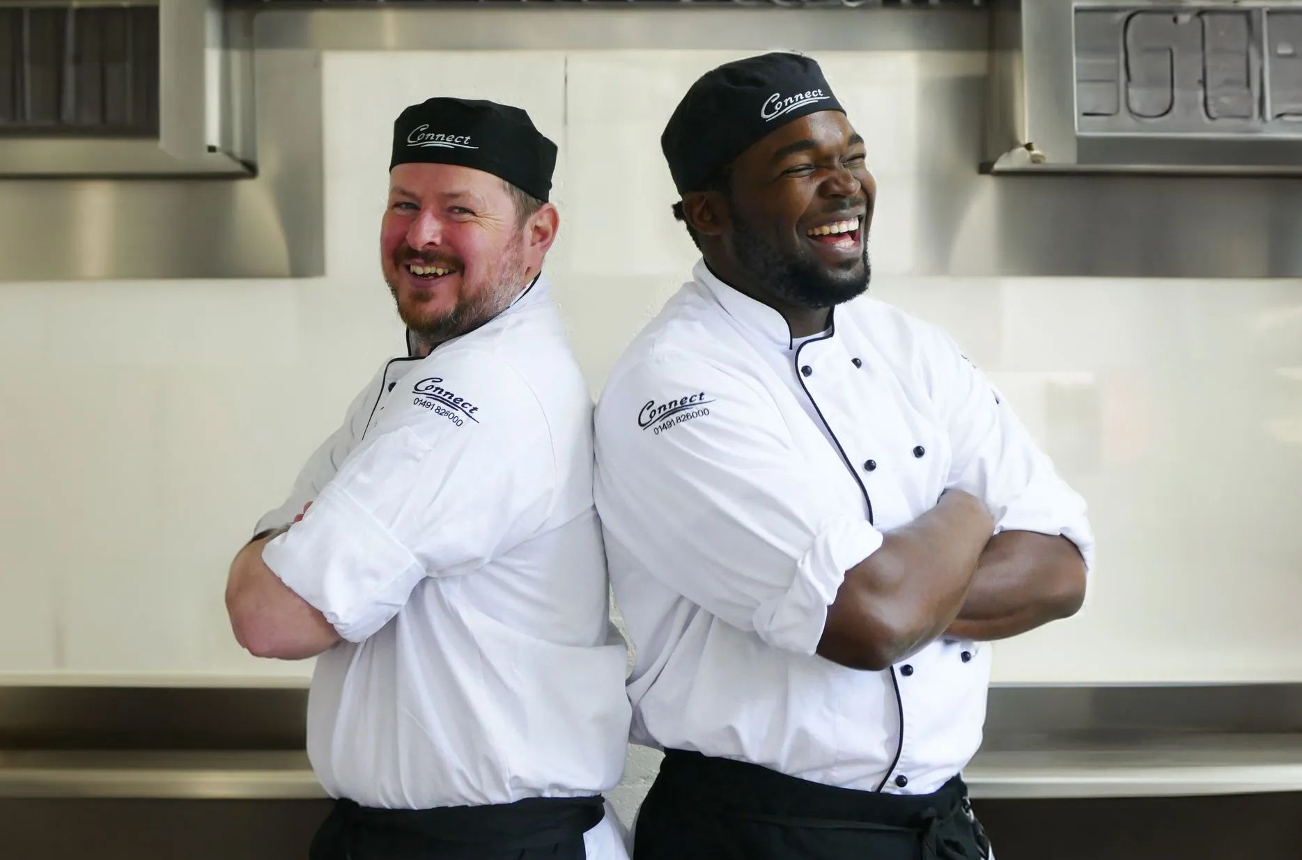 Two male chefs stood back to back laughing in an industrial kitchen, dressed in their chef whites. 