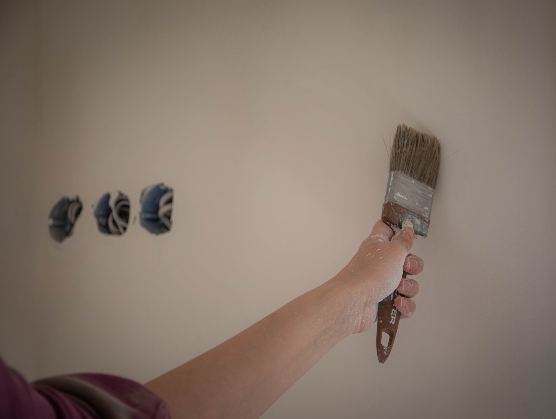 Hand painting beige wall with a brush near three blue electrical outlets