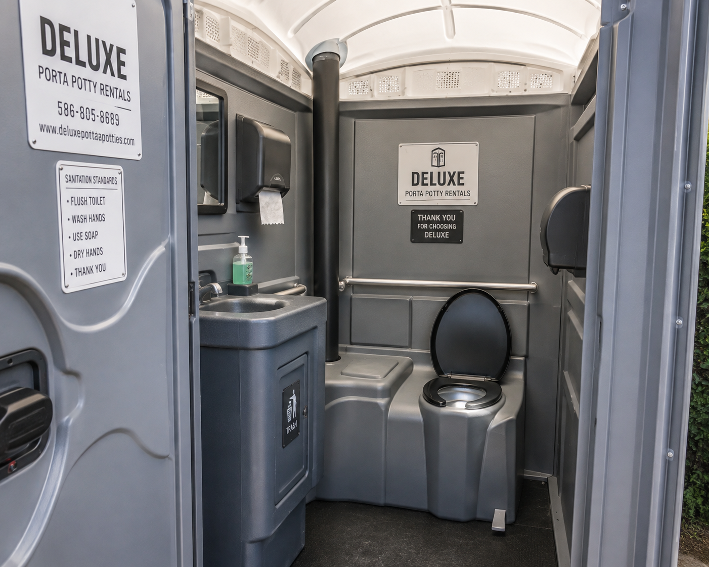 Portable restroom interior with gray walls, toilet, sink, and signage reading “Deluxe.”