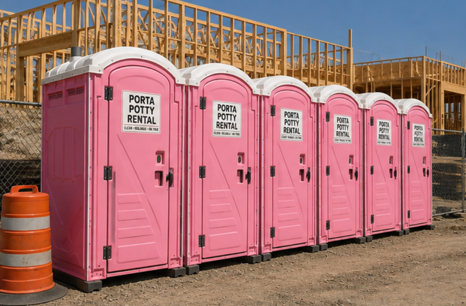 Row of pink portable toilets at a construction site beside an orange barrel and wooden framing